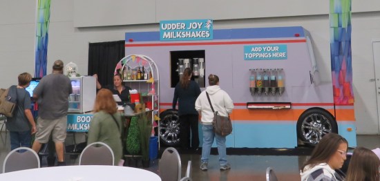 A purple milkshake stand shaped like a food truck, sitting in a convention center with a short line of customers staring at their options.
