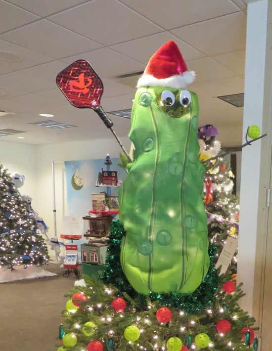 Giant cartoon pickle holds a red flyswatter and sits on a Christmas tree with colored Wiffle balls as ornaments.