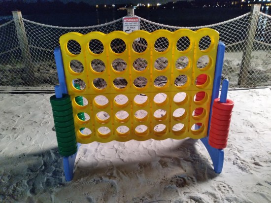 A giant Connect Four game standing on sand. The fence behind it has a sign warning of alligators and snakes.