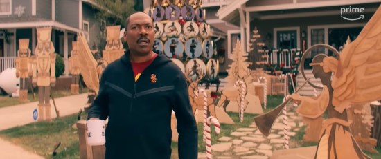 Eddie Murphy as a California dad standing on his lawn covered in big wood-carved Christmas decorations, holding a DAD mug and recoiling at a surprise.
