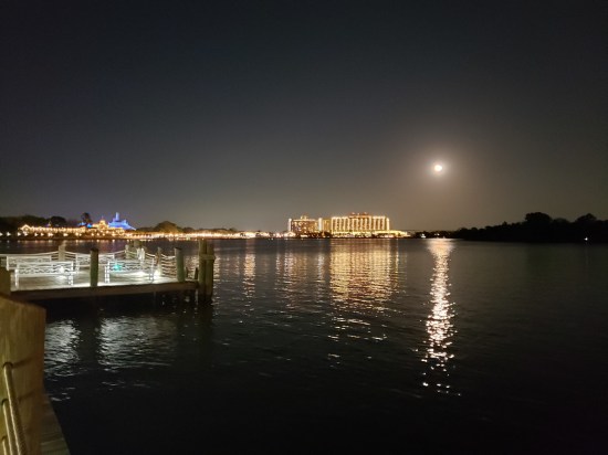 Nighttime shot across a lagoon. On the far shore is a tiny blue castle and a practically neon hotel. The moon shines brightly through a hazy sky, At left is a pier empty but lit.