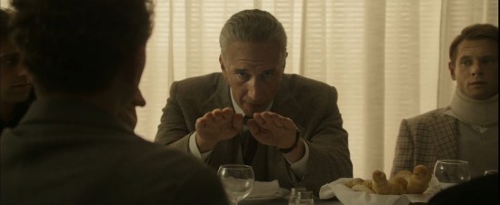 Adam Driver playing the white-haired Enzo Ferrari, sitting at a dinner table with his hands held out palms-down, staring into the camera as he explains what happens when two particular engine parts don't line up perfectly.