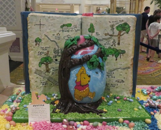 Egg sculpture with Pooh clinging to a helium balloon floating through a forest. Standing behind the egg is a book open to a map of the woods and their trails.