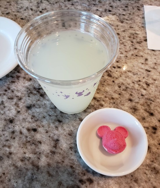 Lemonade in a plastic up. On a tiny plate next to it is a reddish-purple powder tablet shaped like Mickey Mouse's head.