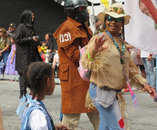 Cheery Black lady in sunglasses and safari explorer clothing, waving at us in a parade. Nearby are cosplayers of Star-Lord, Severus Snape, and Dorothy Gale.