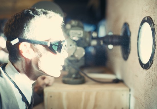 Cillian Murphy as J. Robert Oppenheimer wearing goggles and watching through a tiny bunker window as the first nuclear bomb explodes outside and blinding light comes pouring through.