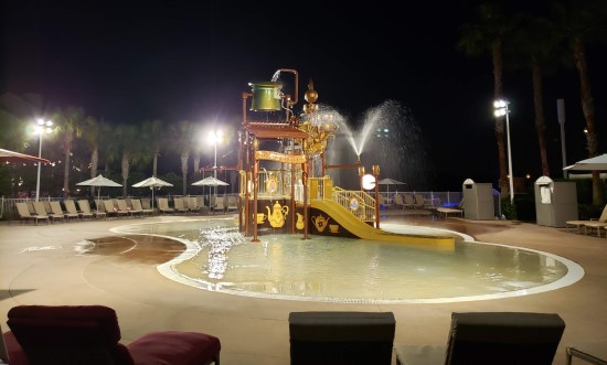 Kid's jungle gym with lots of water-play features installed, lit brightly at night. Water is fountaining but there's no one around.