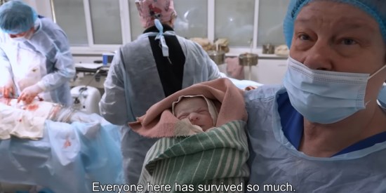 A Ukrainian nurse holds a swaddled, newborn babe. Surgery continues in the background on a patient with bloodied covers.