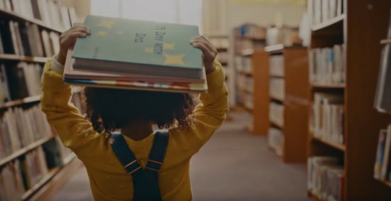 Tiny Black girl carries two books on her head as she walks through a library.