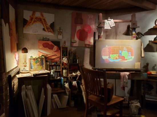 A recreation of a poorly lit artist's studio. Furnishings include a desktop easel, wooden chair, bookshelves, paints, wall art, and unused art canvases.