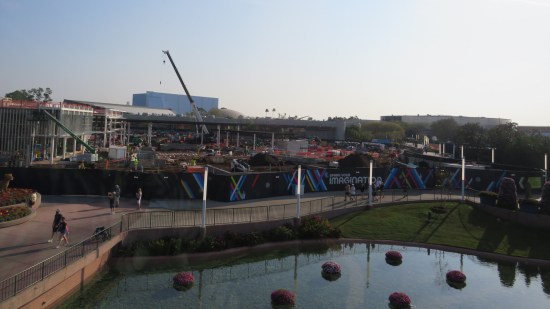 The view from the monorail from the EPCOT park below. Construction site with giant crane, lagoon with flowers in it and a barrier reading "SPARK YOUR IMAGINATION".