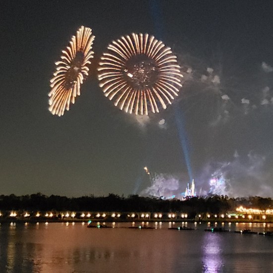 Distant fireworks over water. Two white, flat, titled circles and a single blue spotlight beam shining from the Magic Kingdom.