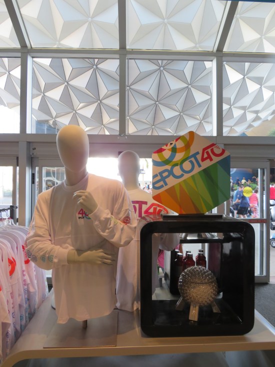 EPCOT souvenir shop next door to Spaceship Earth, which is visible through the store windows. Items for sale include white shirts with pink 40th-anniversary logos and a Spaceship Earth model.