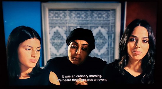 Muslim mother wearing a black cap sits between two adult daughters, one of whom looks away from the camera. Subtitle reads, "It was an ordinary morning. We heard that there was an event."