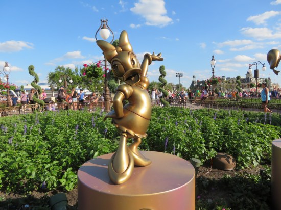 Gilded statue of Daisy Duck, gesturing at the Magic Kingdom. This wide shot shows off more greenery and a few of the tall buildings around the Kingdom.