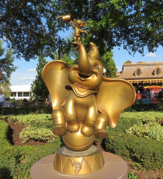 Gilded statue of a baby elephant wearing a hat and balancing on a ball. His trunk holds aloft a mouse dressed like a ringmaster, doffing his hat.