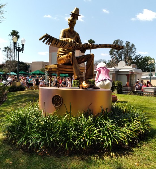 Gilded statue of a jazz musician on a piano bench, playing a magical floating set of 88 keys with no piano attached to them. Behind him is a Disney World "cast member" dressed as Daisy Duck, pantomiming a picnic lunch.