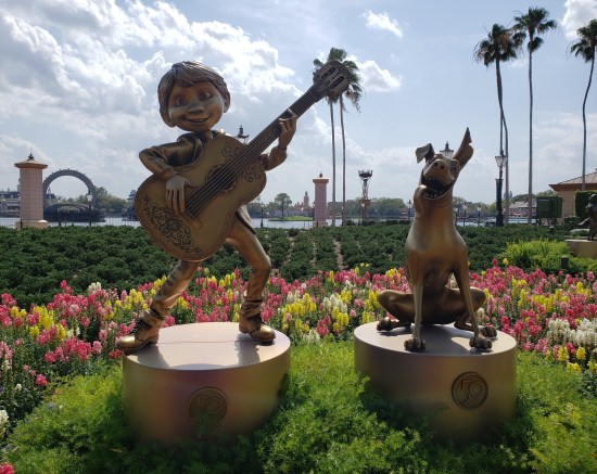 Gold statues of kid mariachi hero and happy ghost dog, in the middle of a garden. A lagoon is in the background.