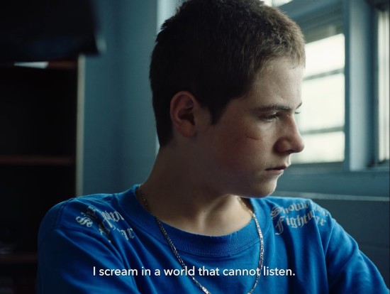 A teenage boy with a scratch on his cheek listens in unease as the offscreen teacher reads his poetry aloud to the class. Subtitle: "I scream in a world that cannot listen."