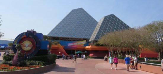 A pair of glass pyramids with oddly extra, short sides near their apexes. Signs call them "Journey Into Imagination With Figment". At far left is a topiary sculpture of a dragon, which will have its own, better photo in the next chapter.