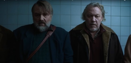 Two older, bearded gents stand in a morgue with people on either side of them. Both are afraid of offending everyone else in the room.