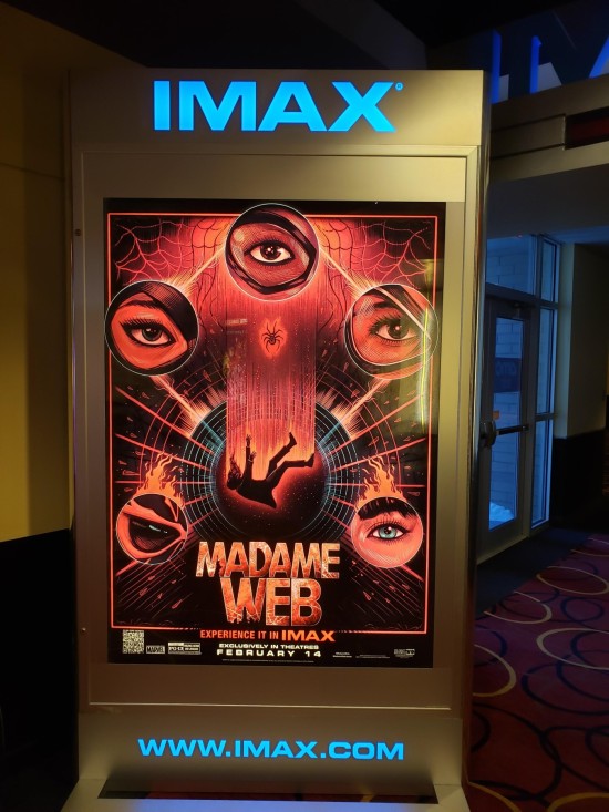 IMAX poster for Madame Web in a theater hallway. Visual elements include five eyes in separate circles surrounding a falling body. In the middle there's a tiny spider. There are concentric circles and some cluttered webbing.