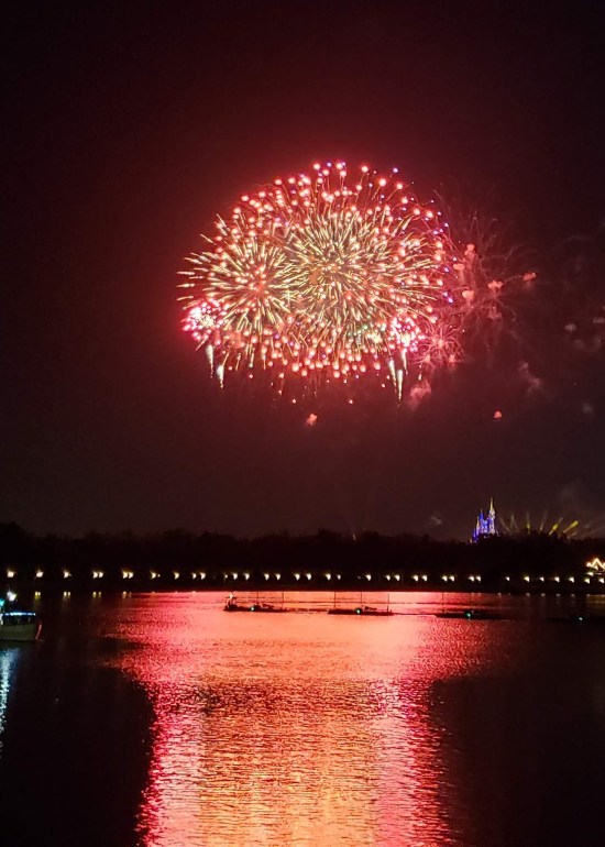 Distant fireworks over water. Huge red ball, great column reflection across the lagoon surface.