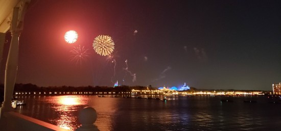 Distant fireworks over water. Medium yellow ball, small red ball. Bright coastal highlights include Tomorrowland in blue and at far right a rectangular hotel. 