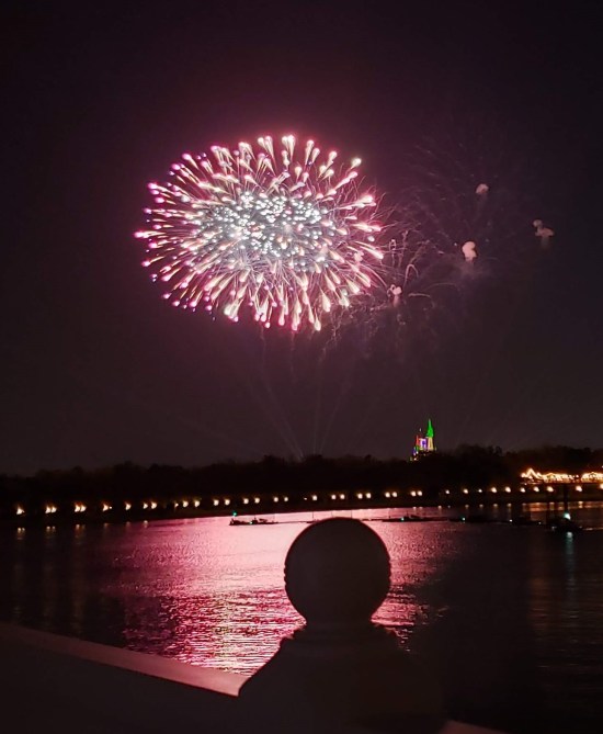 Distant fireworks over water. Overlapping red balls. The ball on the balcony railing obstructs the red water reflections below.