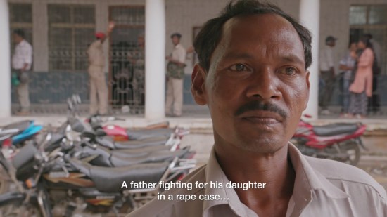 Closeup of a solemn Indian father in front of a courtroom where many motorcycles are parked out front. Subtitles read, "A father fighting for his daughter in a rape case..." and trail off.