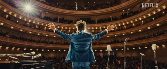 Jon Batiste on stage at Carnegie Hall, viewed from behind as he raises his arms toward an impressed audience.