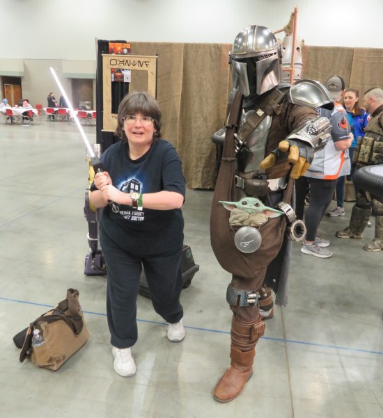 Anne wielding a lightsaber next to a Mandalorian who's trying to use the Force at the camera. Baby Yoda is in his hip pouch.