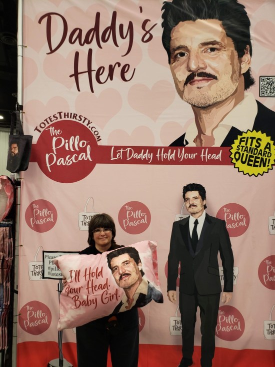 Anne holding a pink pillowcase with Pedro Pascal's face on it and the caption "I'll hold your head, baby girl." Behind her is an ad for same.