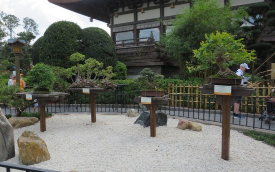 Small enclosure of white sand with four wooden posts, each supporting a tiny platform with a potted bonsai on it.