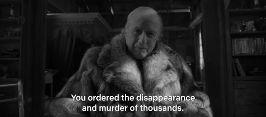Black-and-white shot of an elderly man in a fur coat looking puzzled at the camera. Subtitle from an offscreen accuser reads, "You ordered the disappearance and murder of thousands."