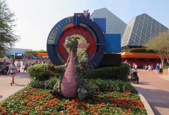 Topiary sculpture of happy, googly-eyed dragon sitting in front of a sign for the "Journey Into Imagination" theater ride.