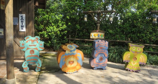 Four parts of a totem pole, each standing separately on a sidewalk next to a wood building.