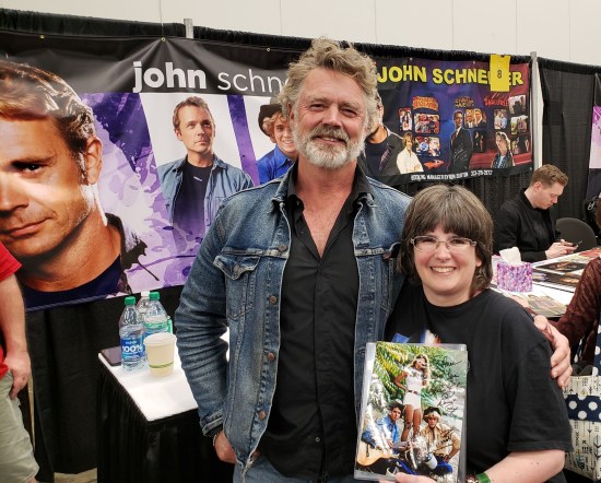 Anne and John Schneider in front of his table. Gray hair, mustache and beard.