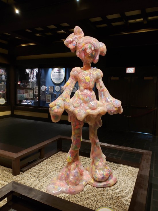 Tall statue of a Harajuku girl with a melting face. The room she's in is poorly lit.