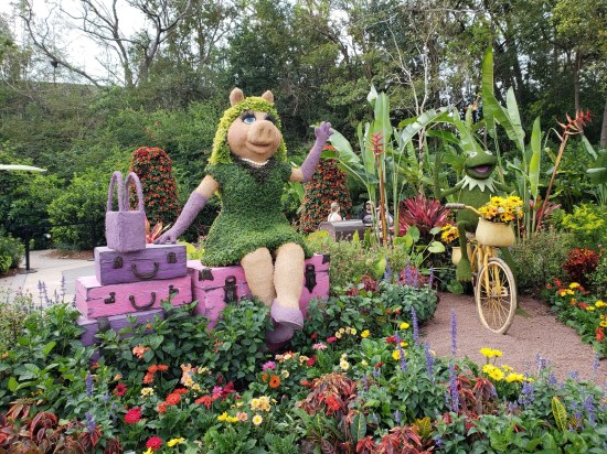 Topiary sculptures of Miss Piggy, who's sitting on several stacked pieces of luggage, and Kermit, riding a bike. Both are waving.
