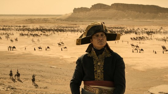 Joaquin Phoenix as Napoleon, scowling and clad in all black including his iconic hat. Far behind him is a desert with some armies milling about and a Western mesa on the horizon.