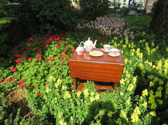 A wooden tea cart with full service laid out, standing in the middle of lots of red, pink, and light green flowers.