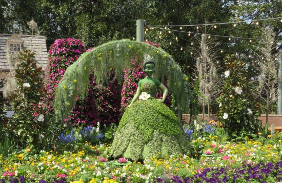 Topiary sculpture of Princess Tiana in a flowerbed with a willow branch arched behind her.