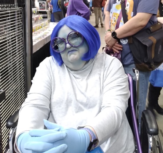 cosplay: Pixar's Sadness. Blue skin makeup, big glasses, large frown, white sweater.