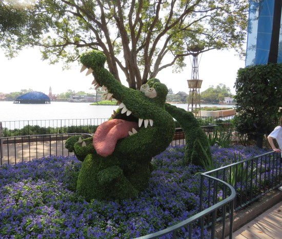Topiary sculpture of cartoon alligator with bulging eyes, jaws open wide and tongue lolling out.