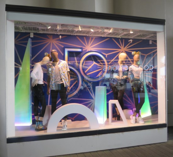 Smartly dressed mannequins in a store window with Disney World's gold 50th anniversary logo prominent in the center.