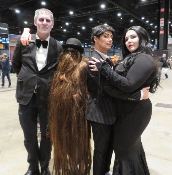 Addams Family cosplayers: Lurch, Cousin It, Gomez and Morticia Addams. The latter two embrace madly.