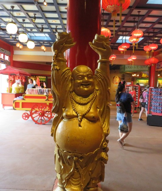 Fake gold statue of Buddha lifting small bowls in each hand over his head. Inside the shop behind him, Chinese lanterns and globe lights line the ceiling.
