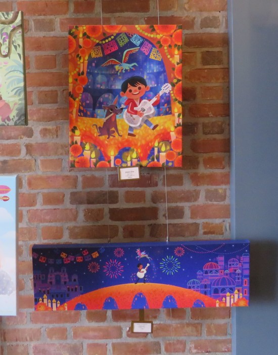 Two paintings resembling concept art from Pixar's "Coco", dangling from strings in front of a brick wall.