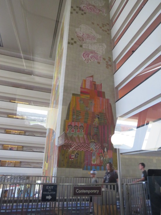 Large, tiled pillar indoor, shot at an upward angle from inside the monorail. Painted on it is a Mexican village and stylized pink clouds.